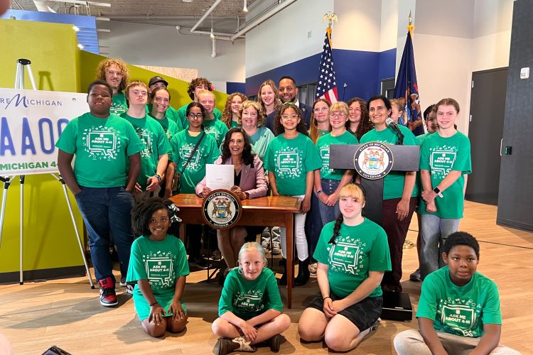 A group of youth in green tshirts standing around Governor Whitmer at a desk.