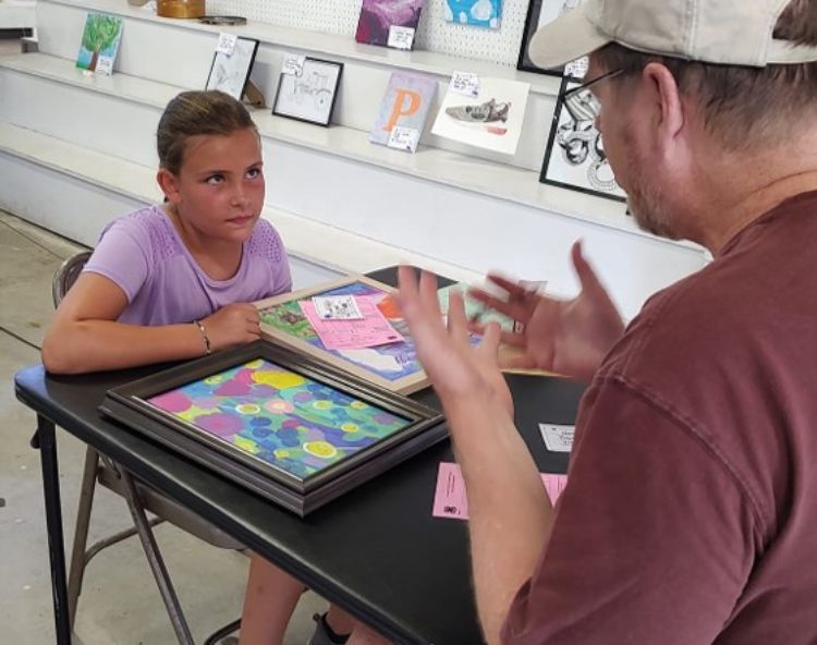 A young girl in a purple shirt sitting with her artwork while talking to an adult.